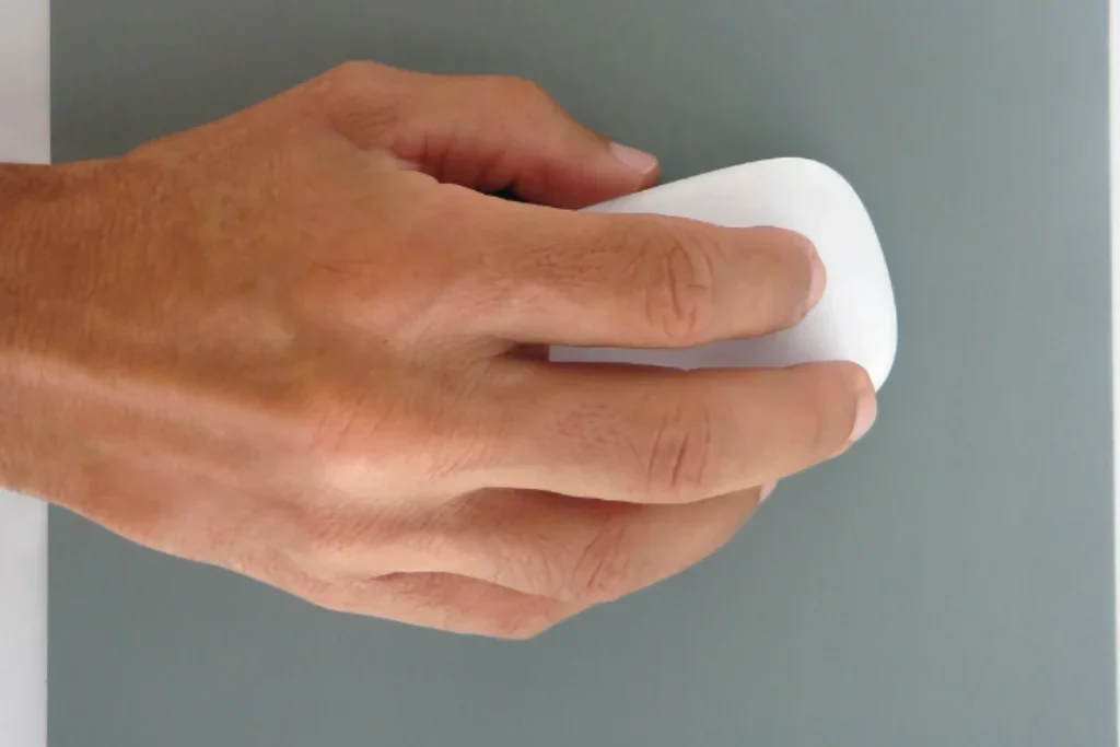 Uma mão segurando um mouse branco sem fio e em cima um mouse pad cinza.
