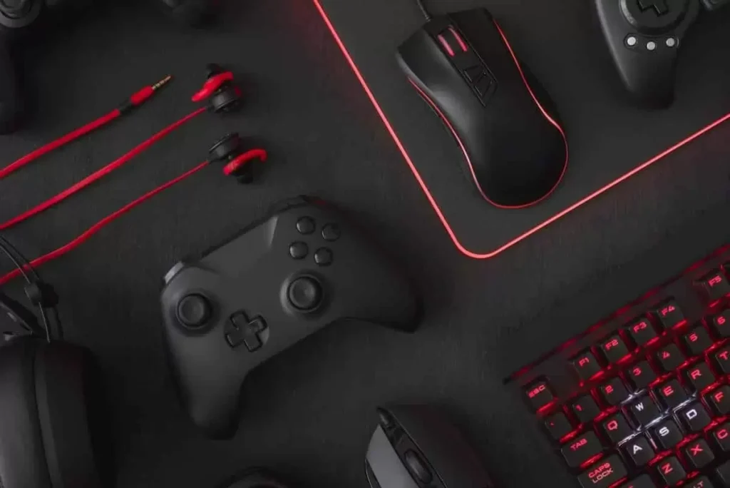 Mouse pad com um controle de xbox em cima, um fone de ouvido pequeno, um dois mouses pretos, um teclado e com uma ilustração em vermelho e preto.