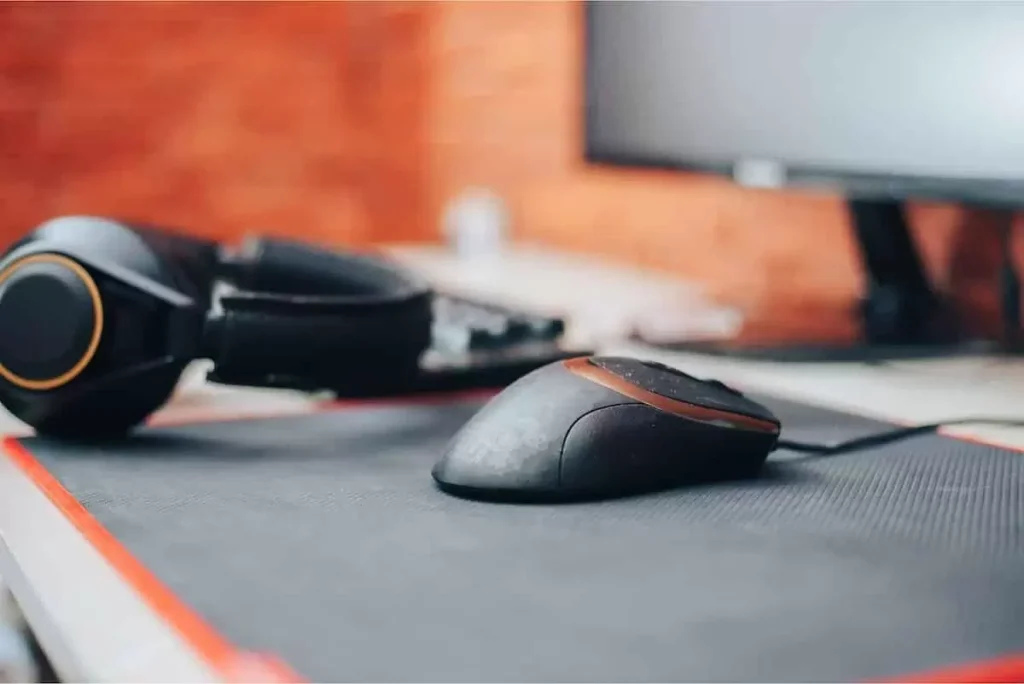 Um mouse em cima de um mouse pad e de fundo um fone de ouvido e um monitor de PC.